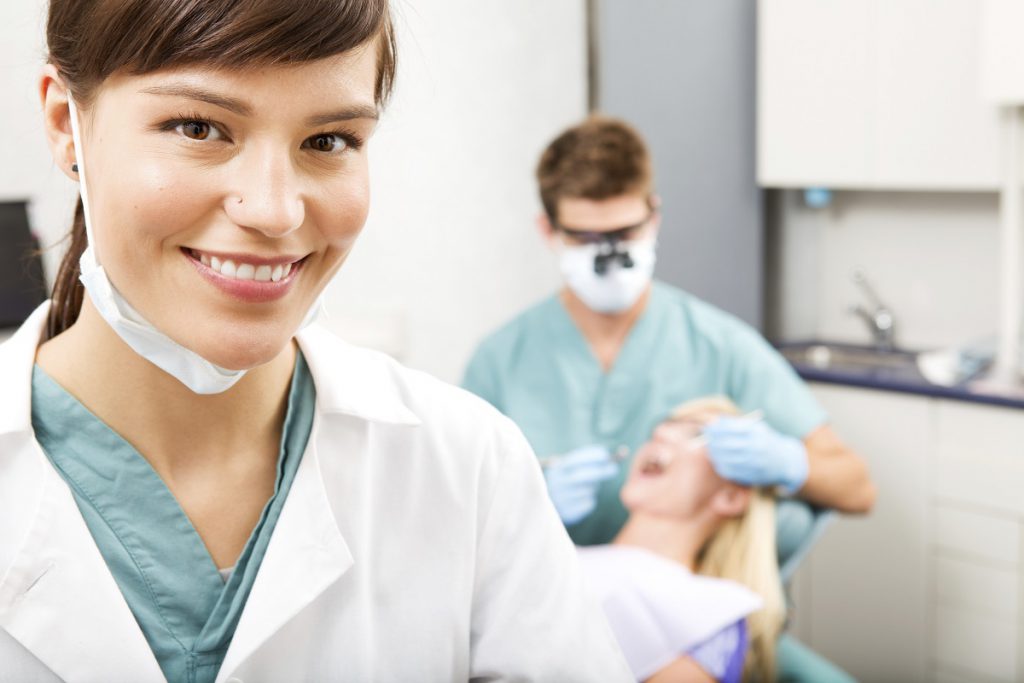 A dentist works on a patient in the background while his dental assistant smiles